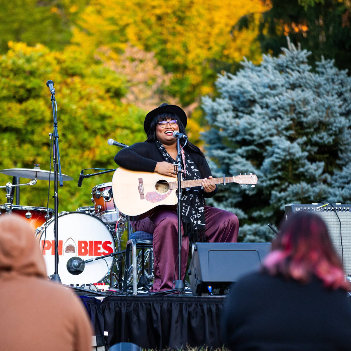 A student sitting in a chair, singing into a mic, and playing guitar in the arboretum